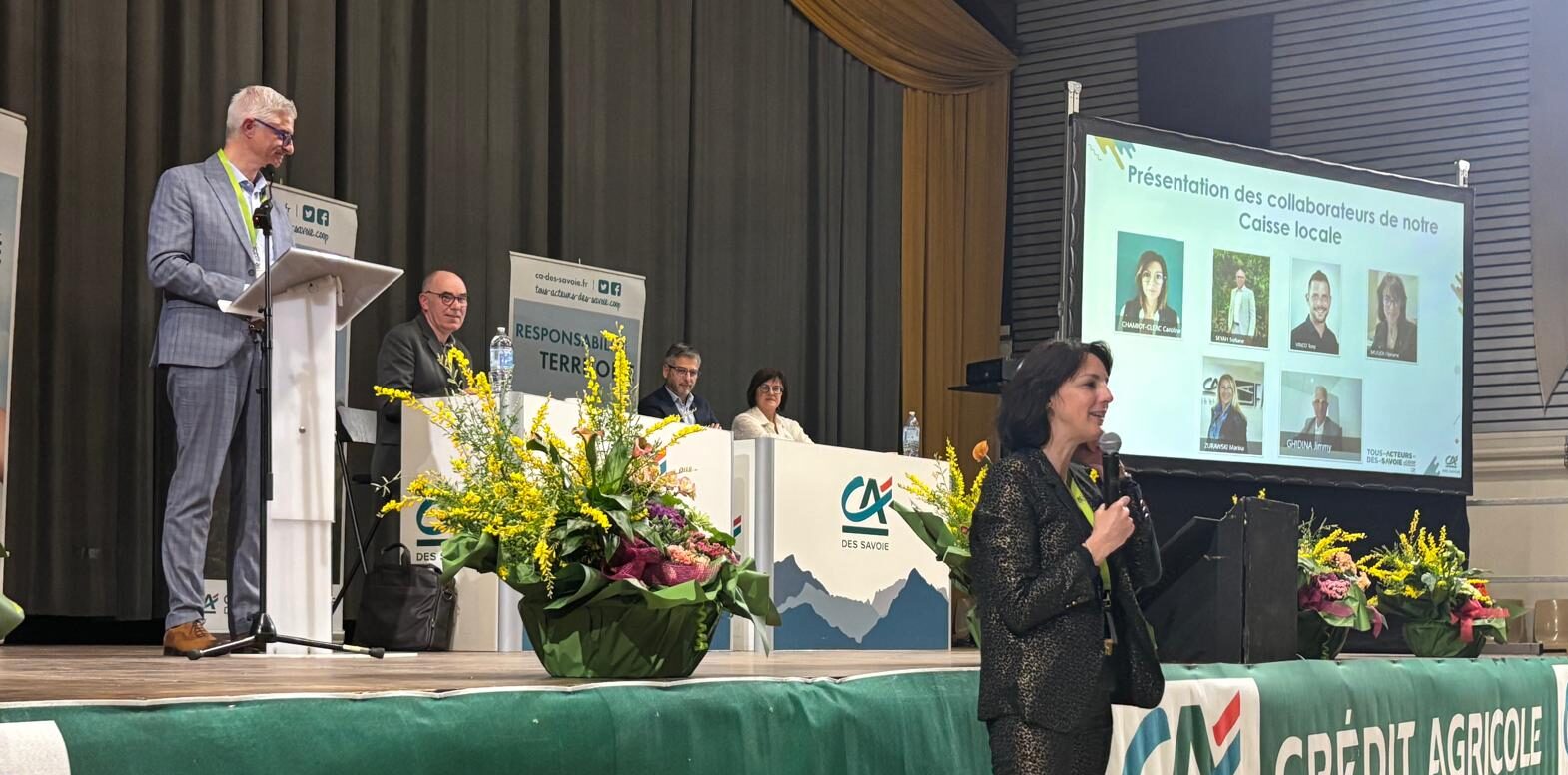 Sur scène 4 personnes assisent à un bureau. et devant la scène une personne faisant un discours.