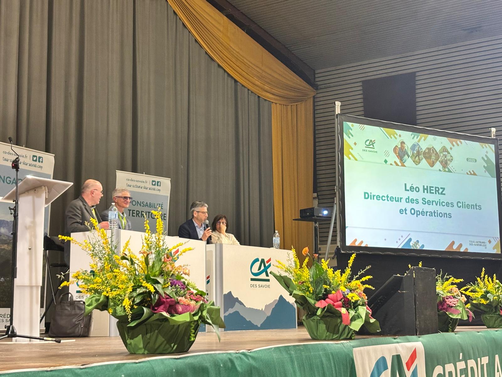 Sur scène 4 personnes assise à des bureaux, l'une parant dans un micro et un power point est diffusé. Des fleurs sur le devant de la scène pour décorer.
