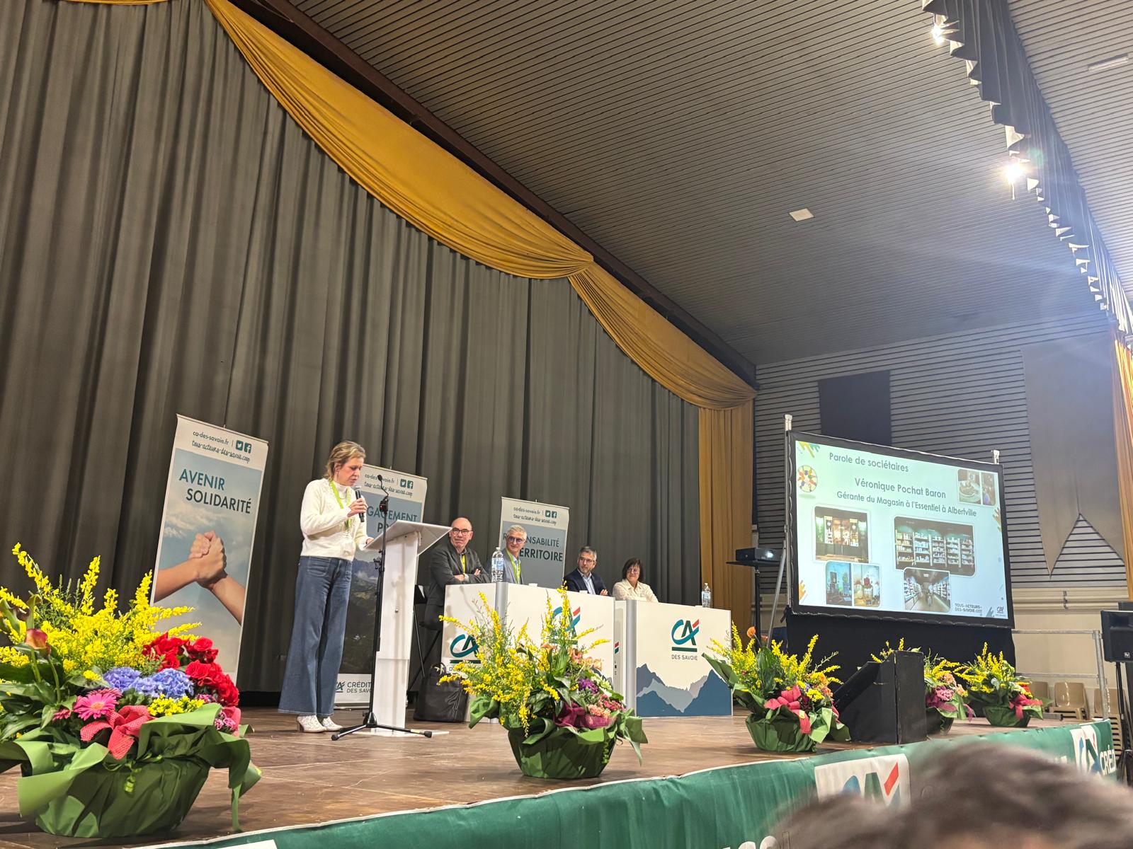 Sur scène, une femme faisant un discours, 4 personnes assisent à un bureau.