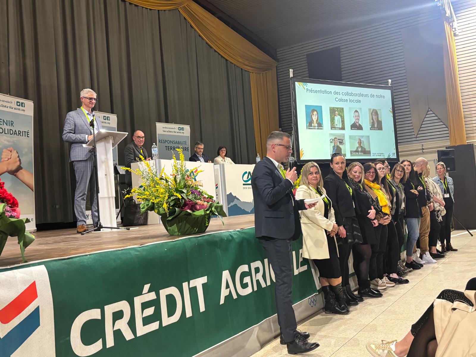 Sur scène, 4 personnes assise à des bureaux et un power point est diffusé. Devant la scène plusieurs personnes debout face au public. Des fleurs sur le devant de la scène pour décorer.