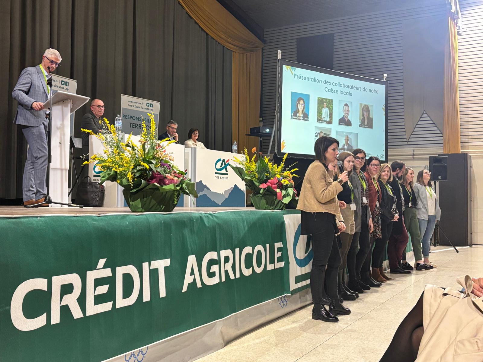 Sur scène, 4 personnes assise à des bureaux et un power point est diffusé. Devant la scène plusieurs personnes debout face au public. Des fleurs sur le devant de la scène pour décorer.